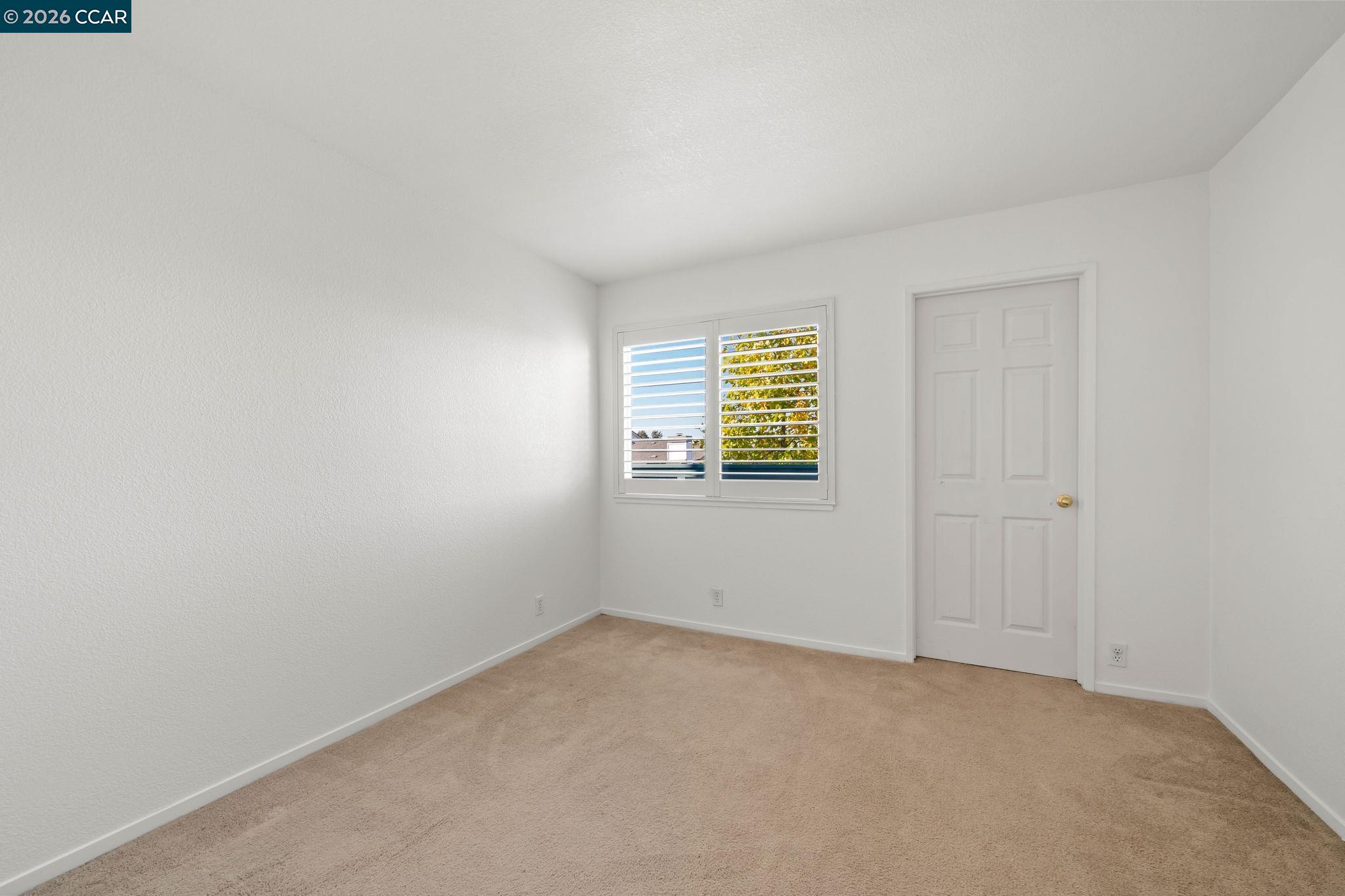 Empty carpeted room with white walls, plantation shutters, and a paneled door.
