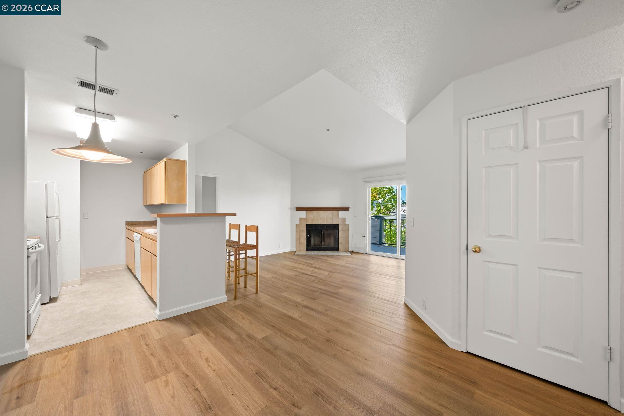 Open-plan kitchen and living room with light wood floors, bar seating, a tiled fireplace, and balcony access.