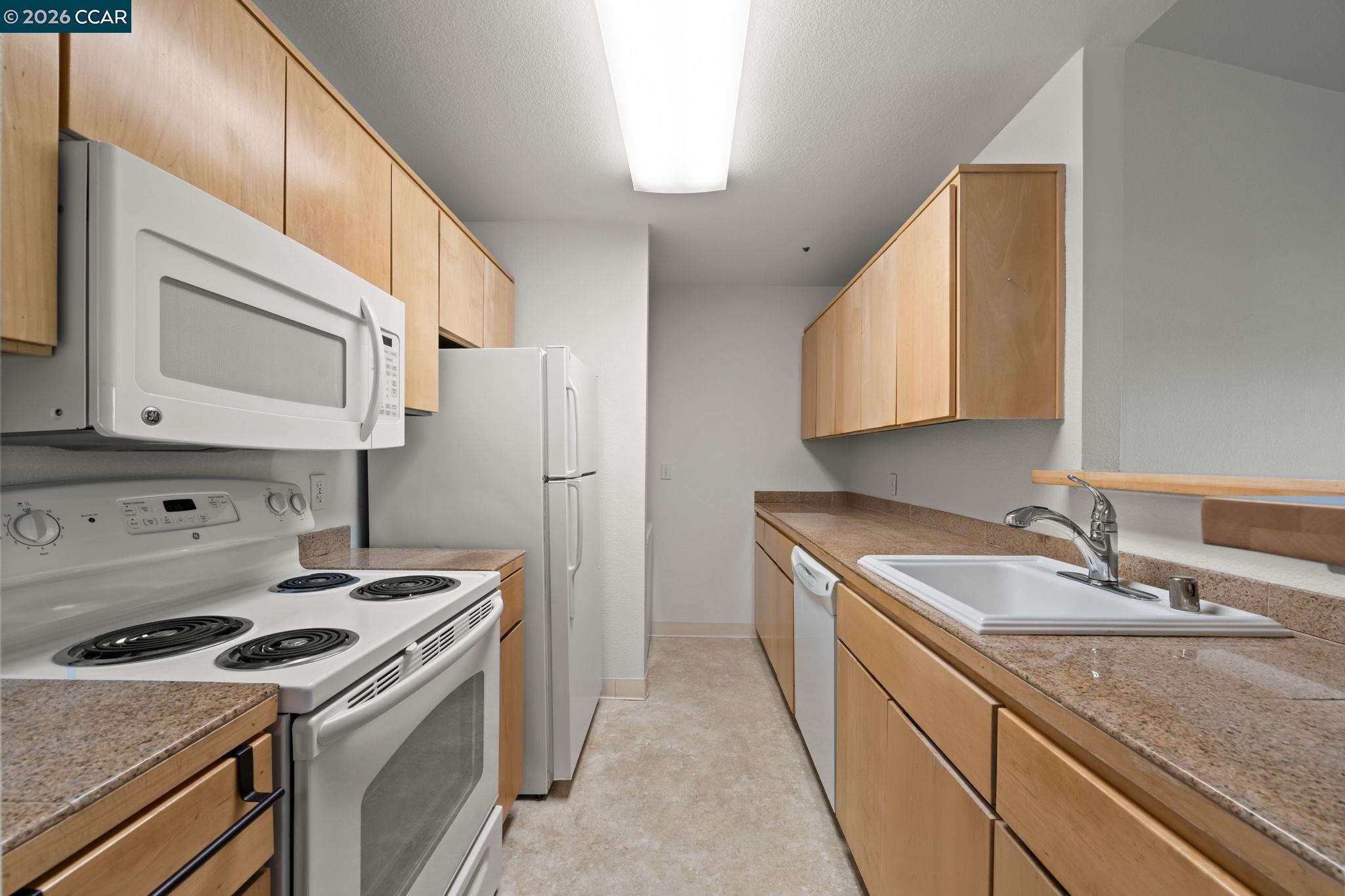Kitchen with light wood cabinets, speckled countertops, and white appliances.