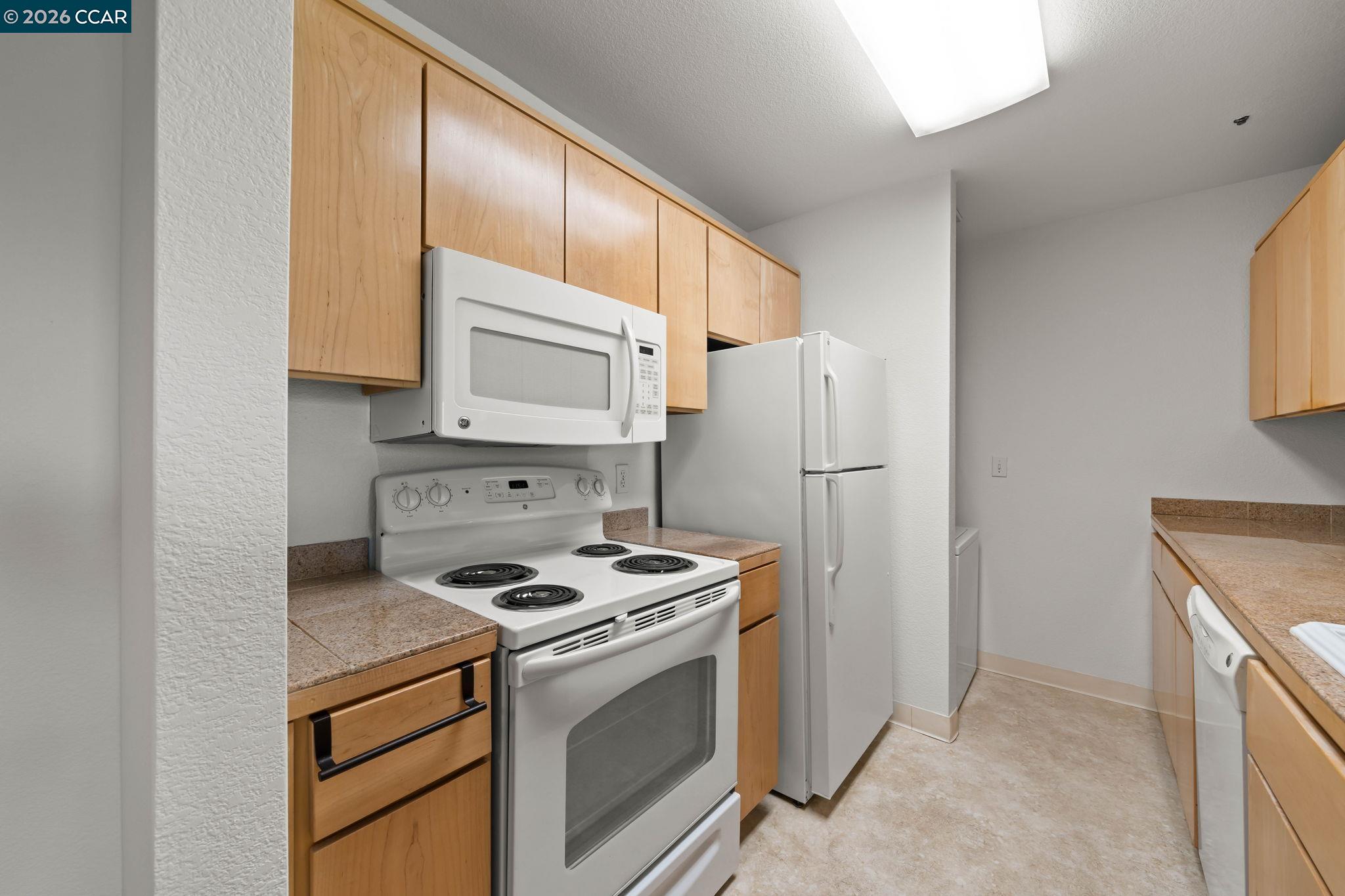Kitchen with light wood cabinets, speckled counters, and white appliances.
