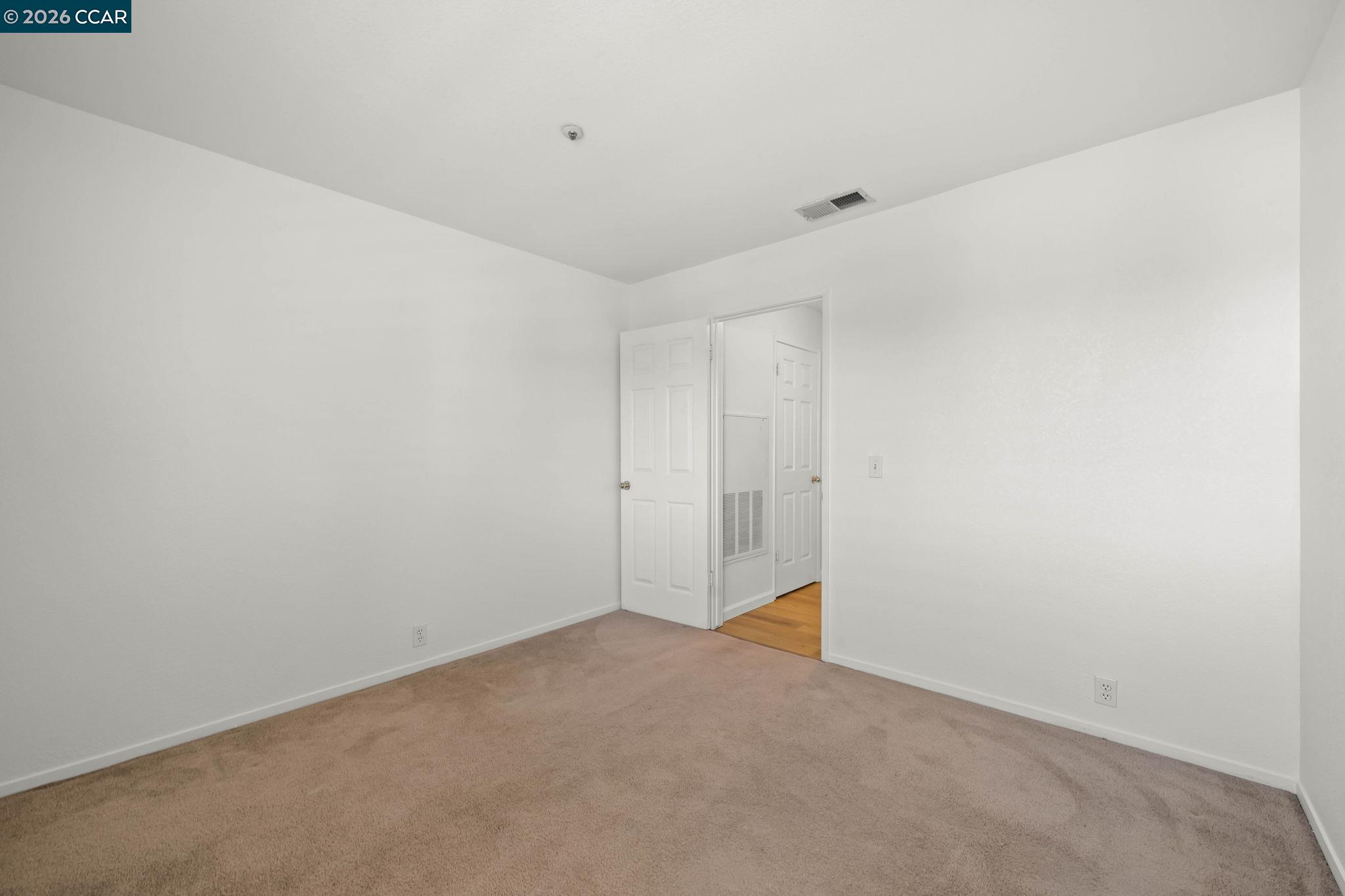 Empty bedroom with beige carpeting and an open doorway to a wood-floored area.