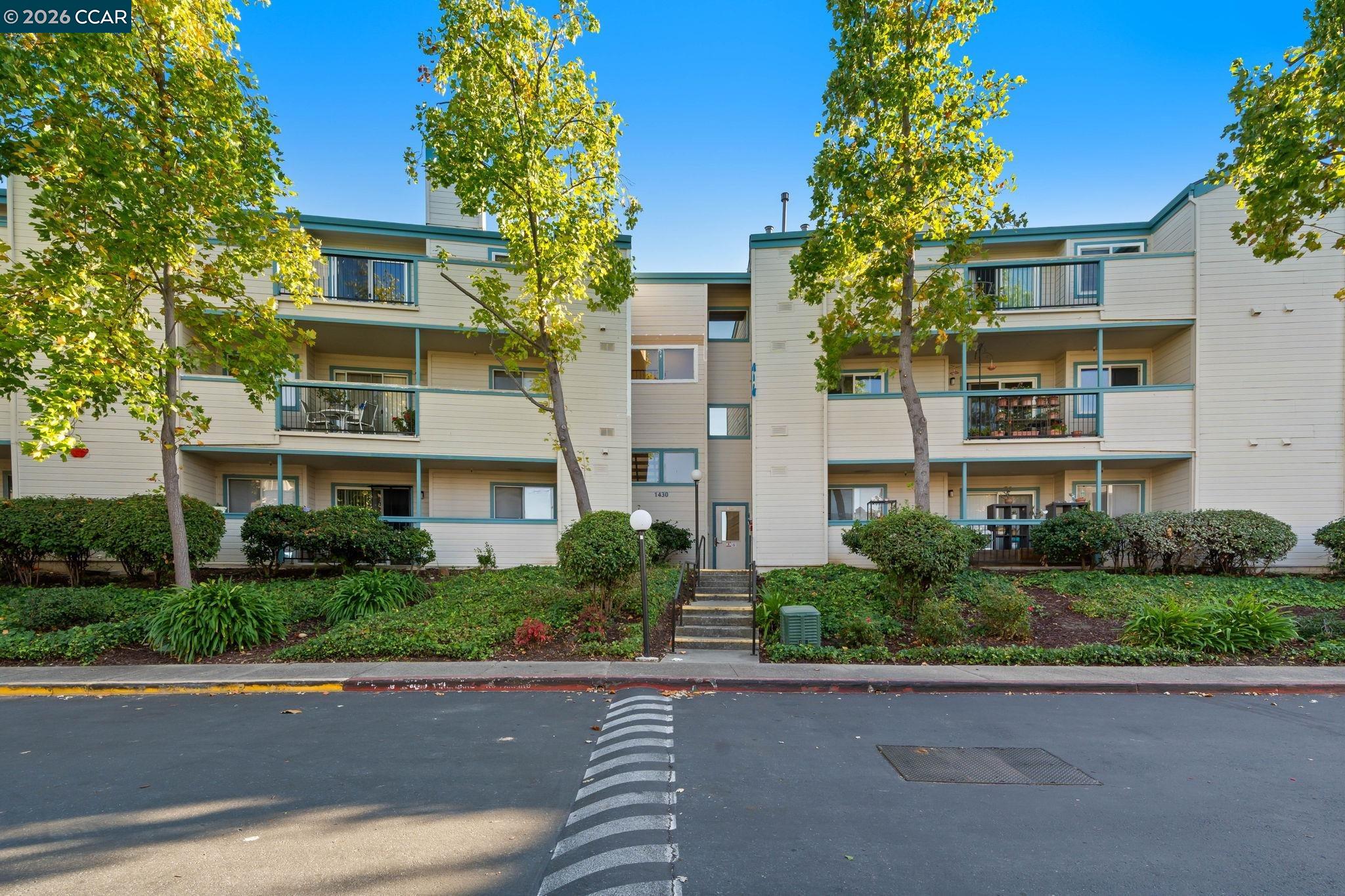 Three-story condominium building with teal balconies, mature trees, and landscaped shrubs along the front.