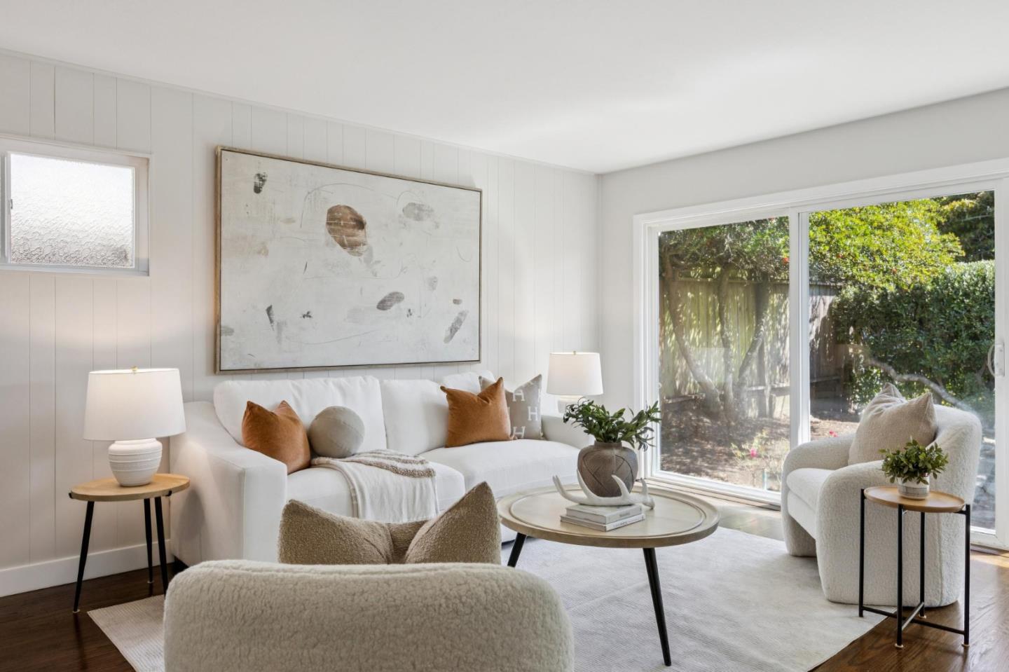 Bright living room with white seating, abstract wall art, and sliding glass doors to a leafy yard.