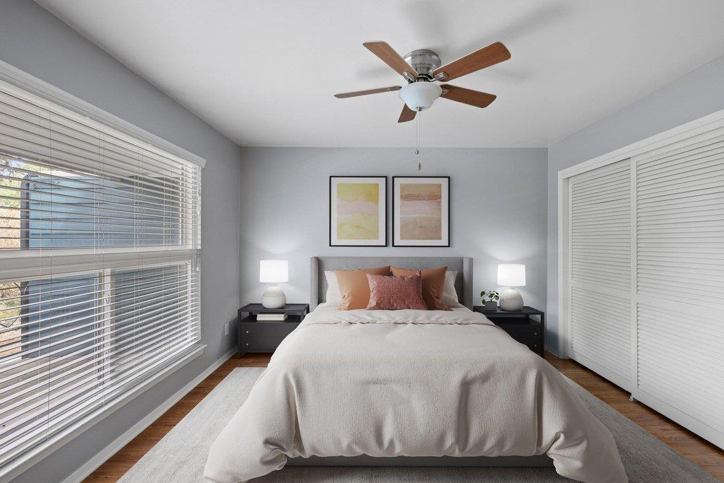 Bedroom with a neatly made bed, blush pillows, ceiling fan, and wide windows with blinds.