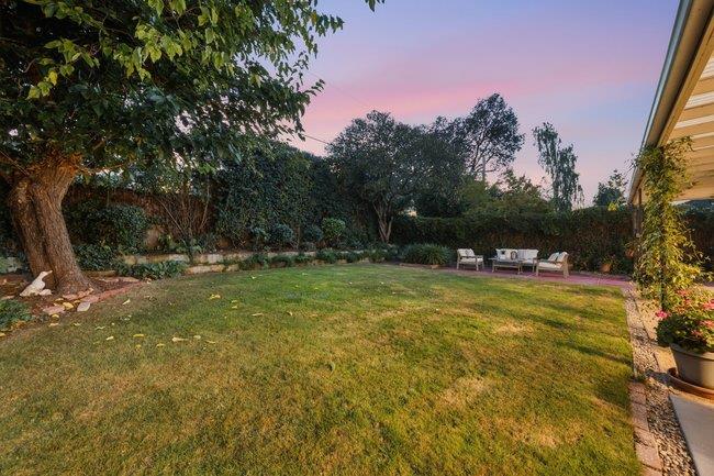 Backyard lawn bordered by mature trees, with patio seating beneath a pink dusk sky.