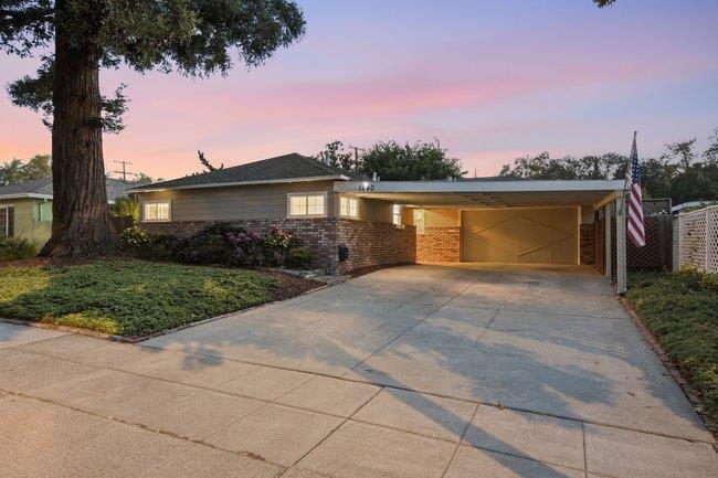 Single-story home with an attached two-car garage, wide driveway, mature tree, and landscaped front garden at dusk.