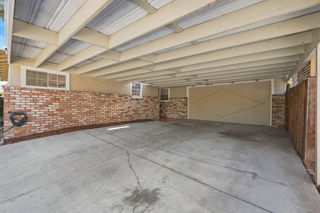Covered carport with exposed beams, brick half-walls, and a concrete parking area.
