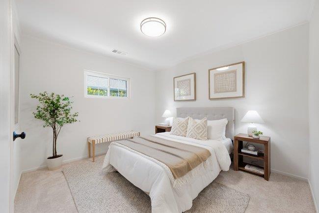 Staged bedroom with a white bed, beige bedding, framed artwork, and a potted tree.