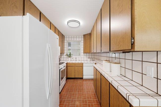 Galley kitchen with wood cabinets, white appliances, and a reddish brick floor.