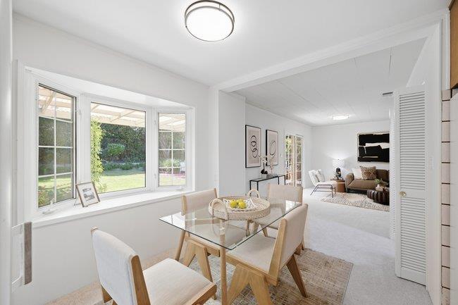 Dining room with a glass-top table beside large windows, opening into a furnished living room.