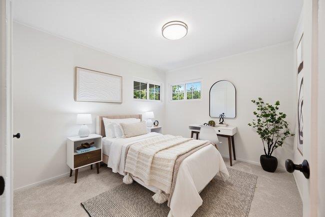 Staged bedroom with a white bed, small windows, a desk and mirror, and a potted plant.