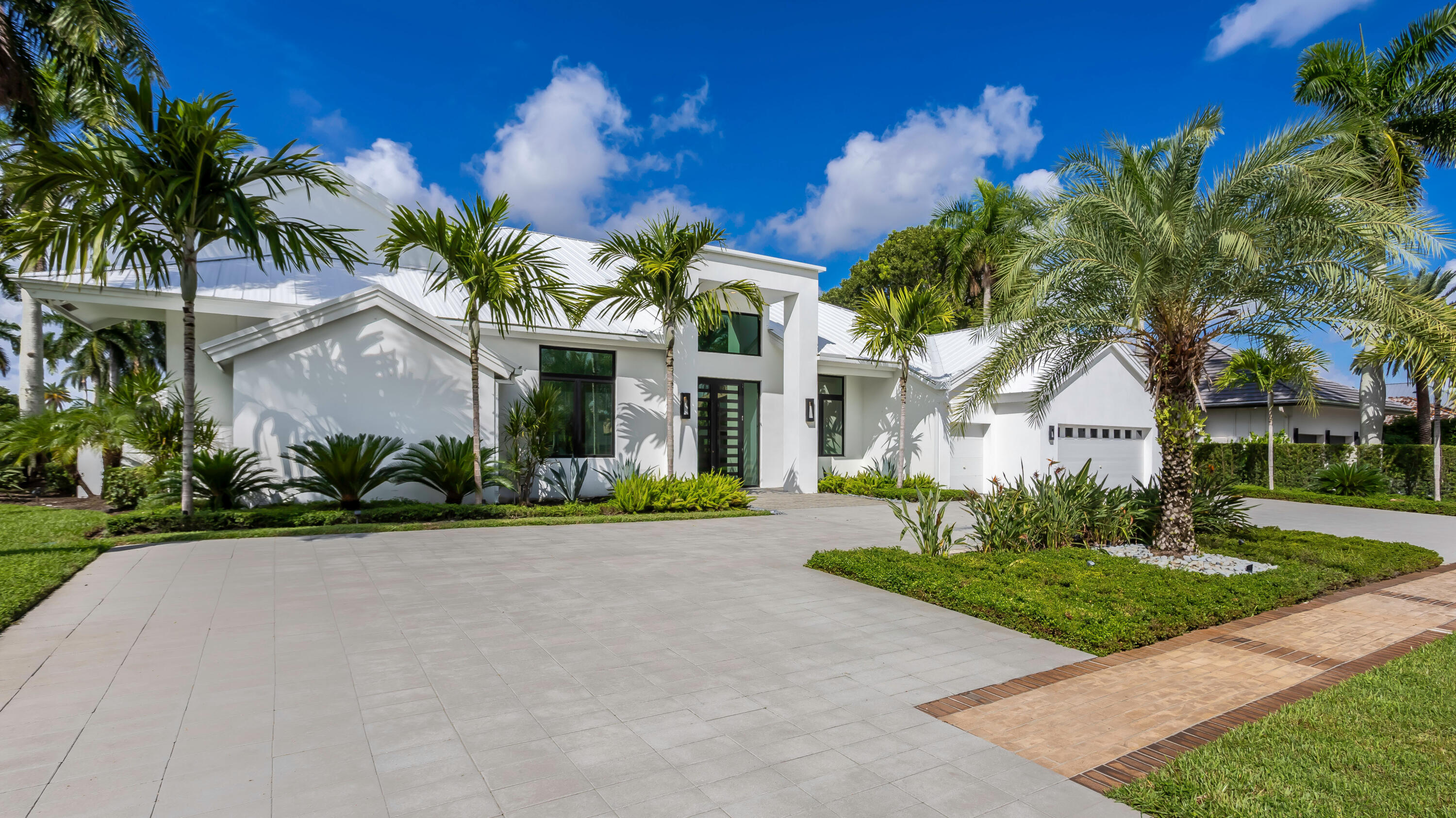 Modern white home with a metal roof, palm trees, and a broad paved driveway.