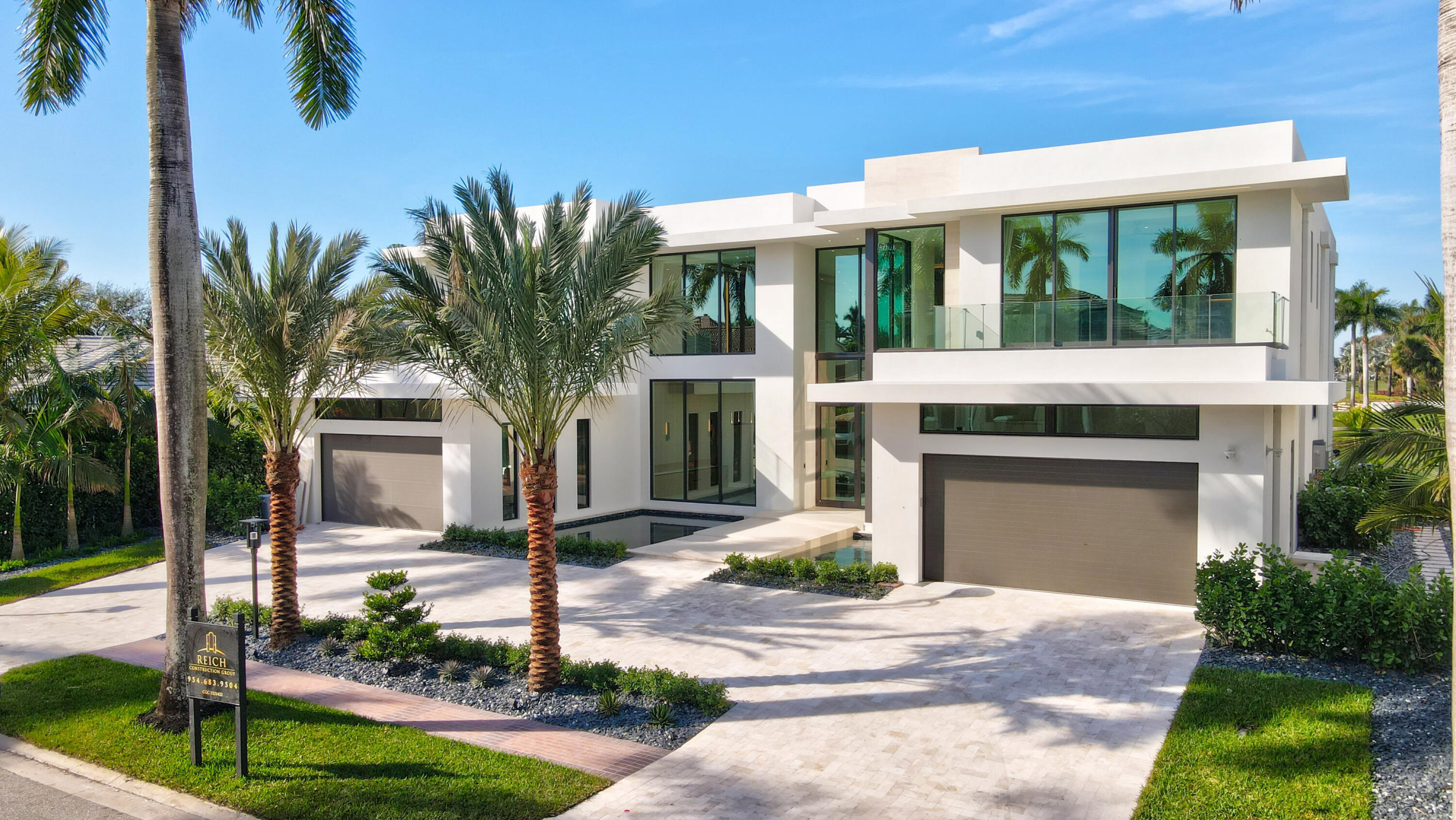 Modern white two-story home with glass balconies, broad driveway, and palm trees.