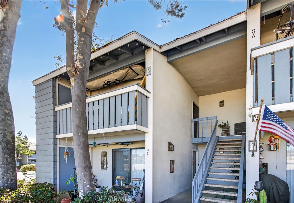 Two-story condominium exterior with blue-gray balconies, an outdoor staircase, and a flag beside the entry.