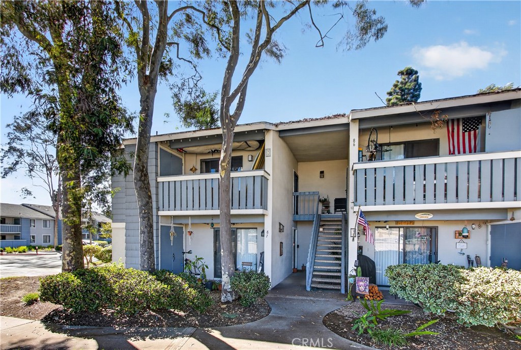 Two-story condominium exterior with balconies, a central staircase, mature trees, and American flags.