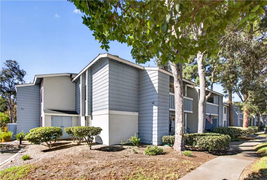 Blue-gray attached condominium building with landscaped shrubs and mature trees along the sidewalk.