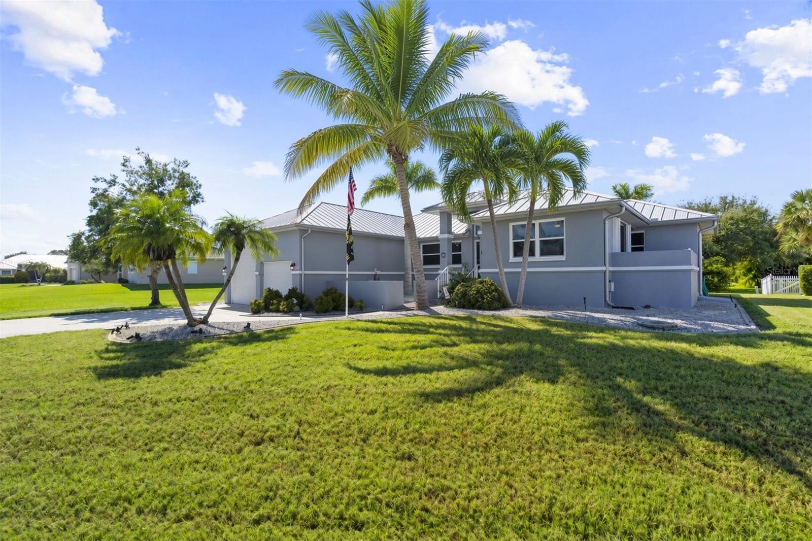 Gray single-story home with a metal roof, tall palms, and a broad green lawn.