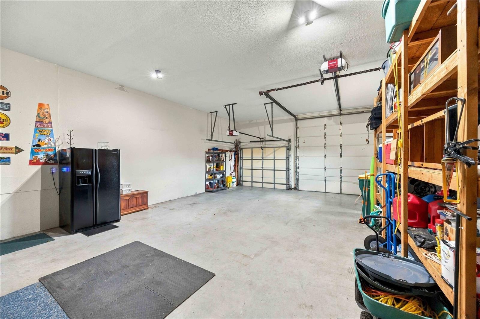 Spacious garage with two garage doors, a refrigerator, and storage shelves along the right wall.