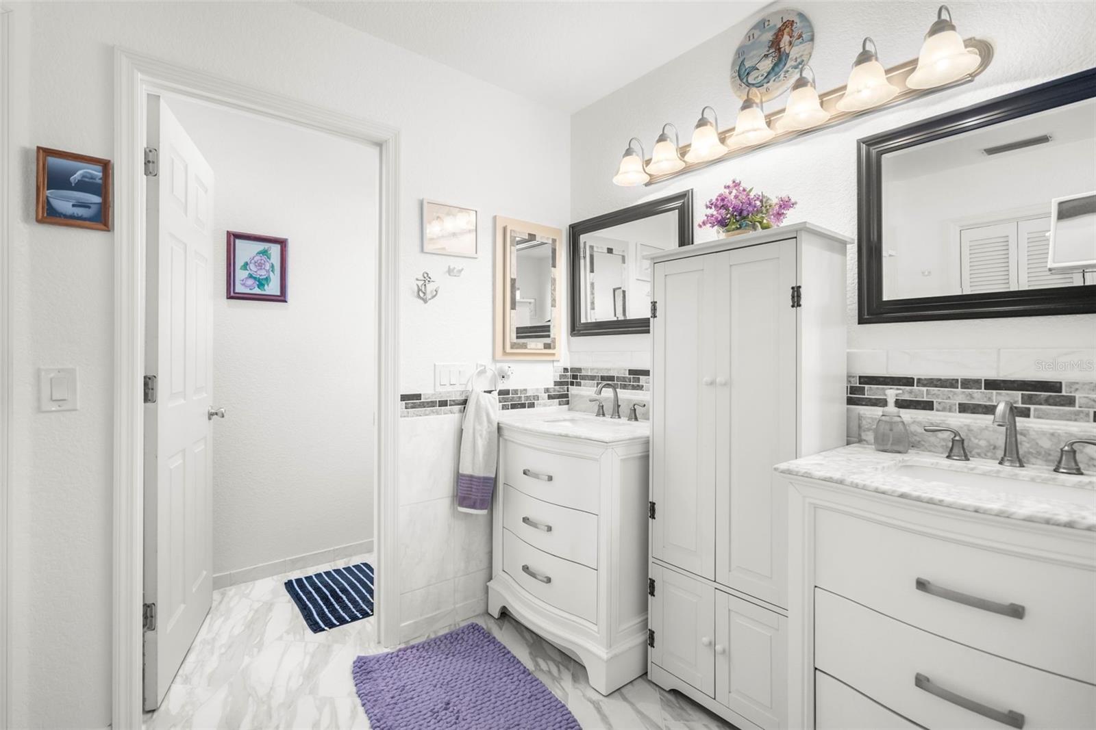 Bright bathroom with white double vanity, marble-look counters, tiled floor, and purple rugs.