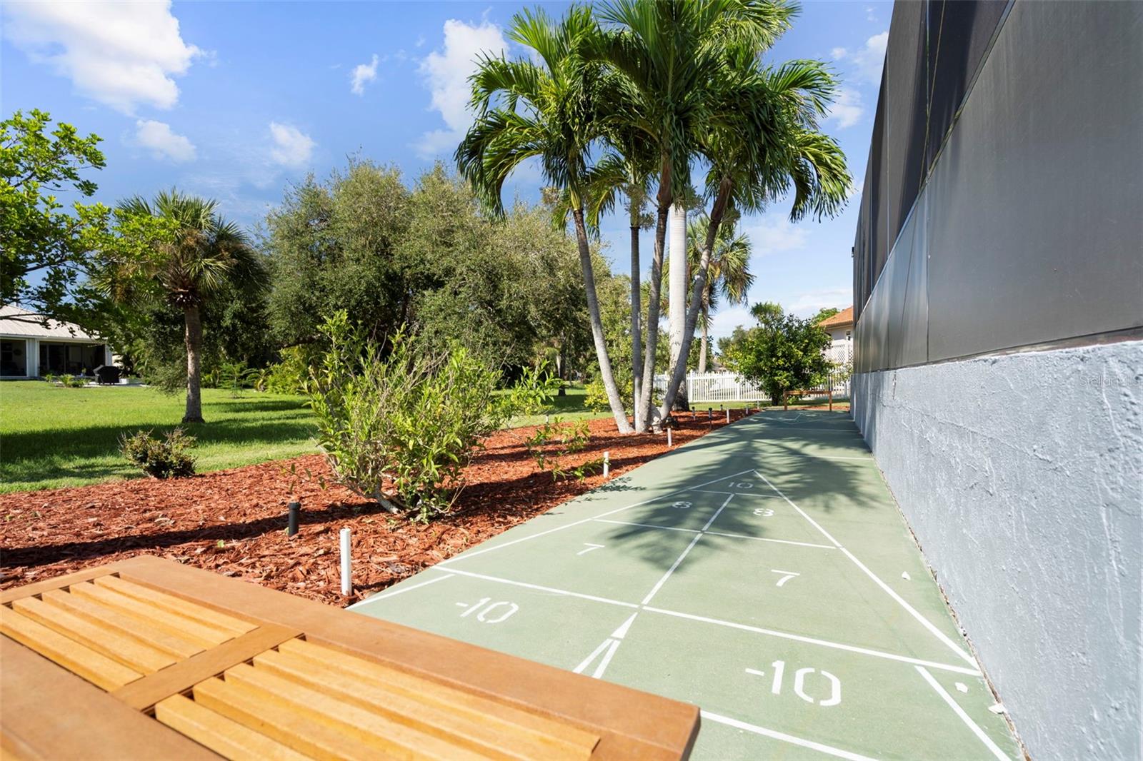 Outdoor shuffleboard court beside a building, bordered by palm trees and landscaped grounds.