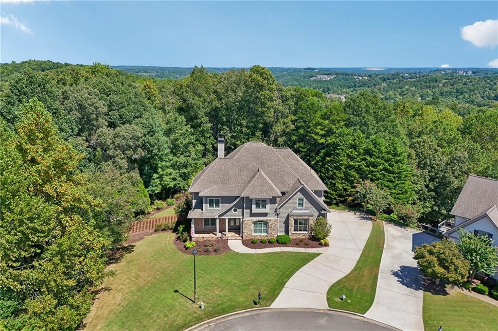 Aerial view of a two-story stone-and-siding house with a curved driveway and wooded surroundings.