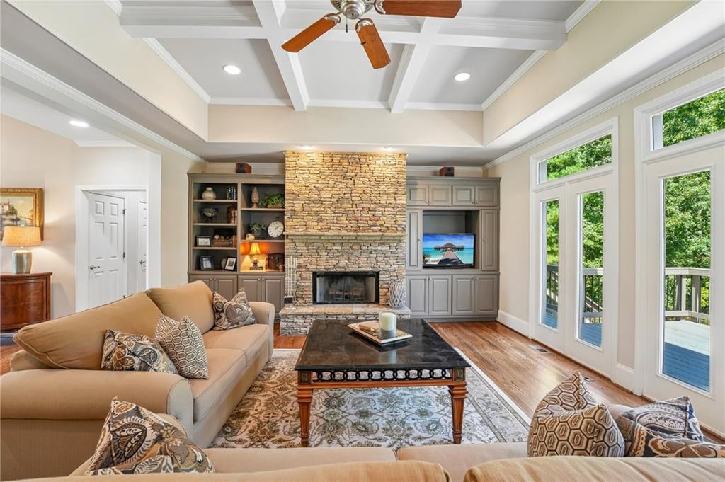 Living room with a stone fireplace, coffered ceiling, built-ins, and tall windows overlooking greenery.