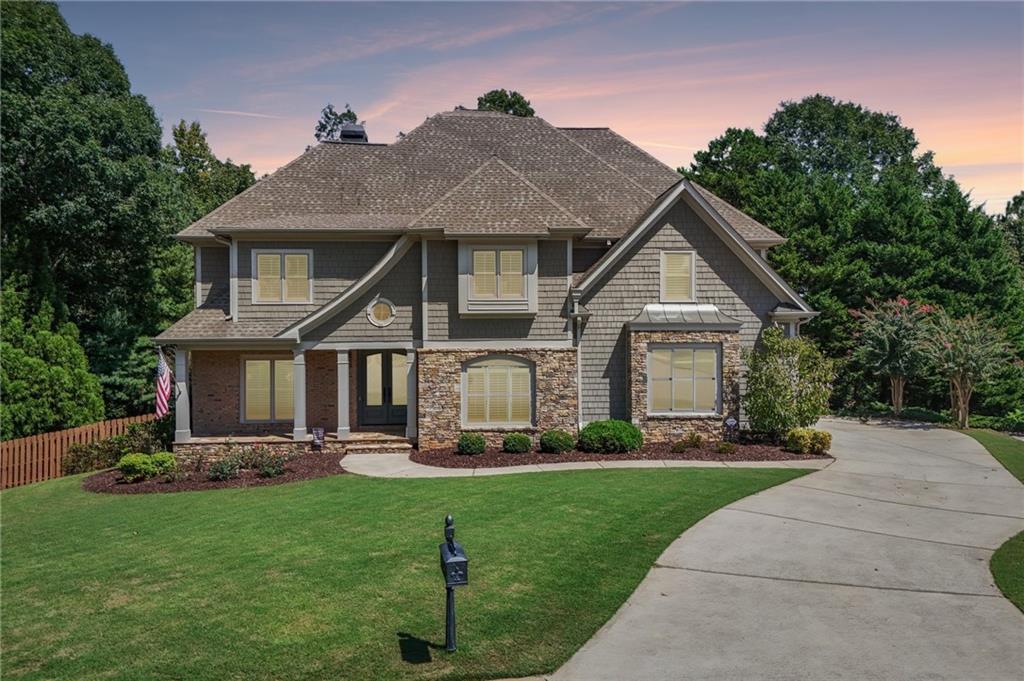 Two-story gray-shingled house with stone accents, landscaped lawn, and curved driveway at dusk.