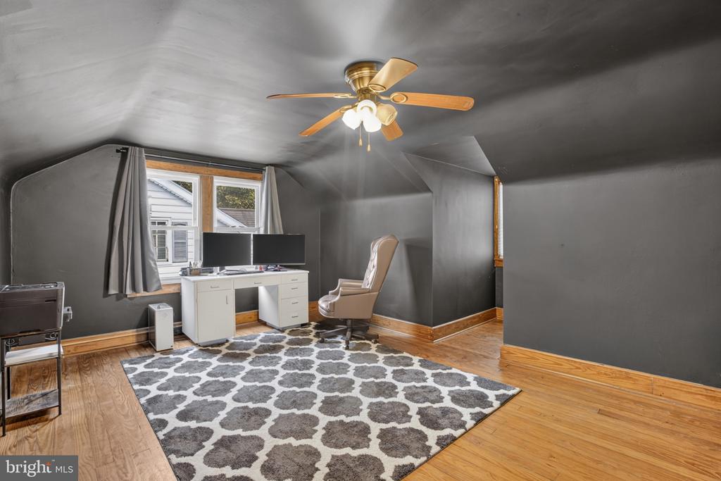 Attic office with a white desk, two monitors, patterned rug, and ceiling fan.
