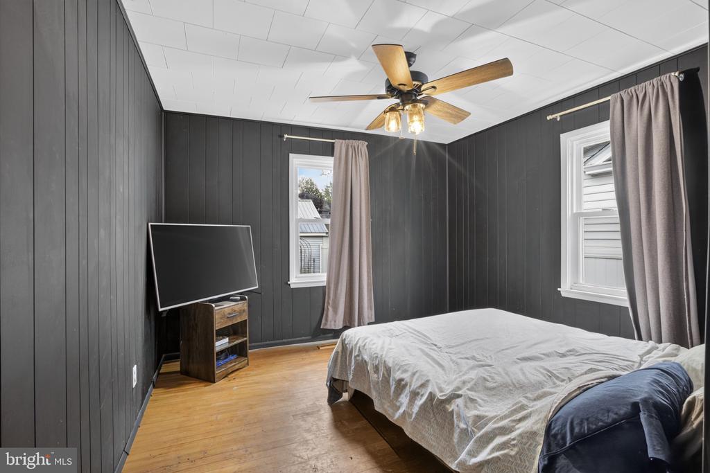 Bedroom with charcoal paneled walls, hardwood floors, a ceiling fan, and a bed beneath two curtained windows.