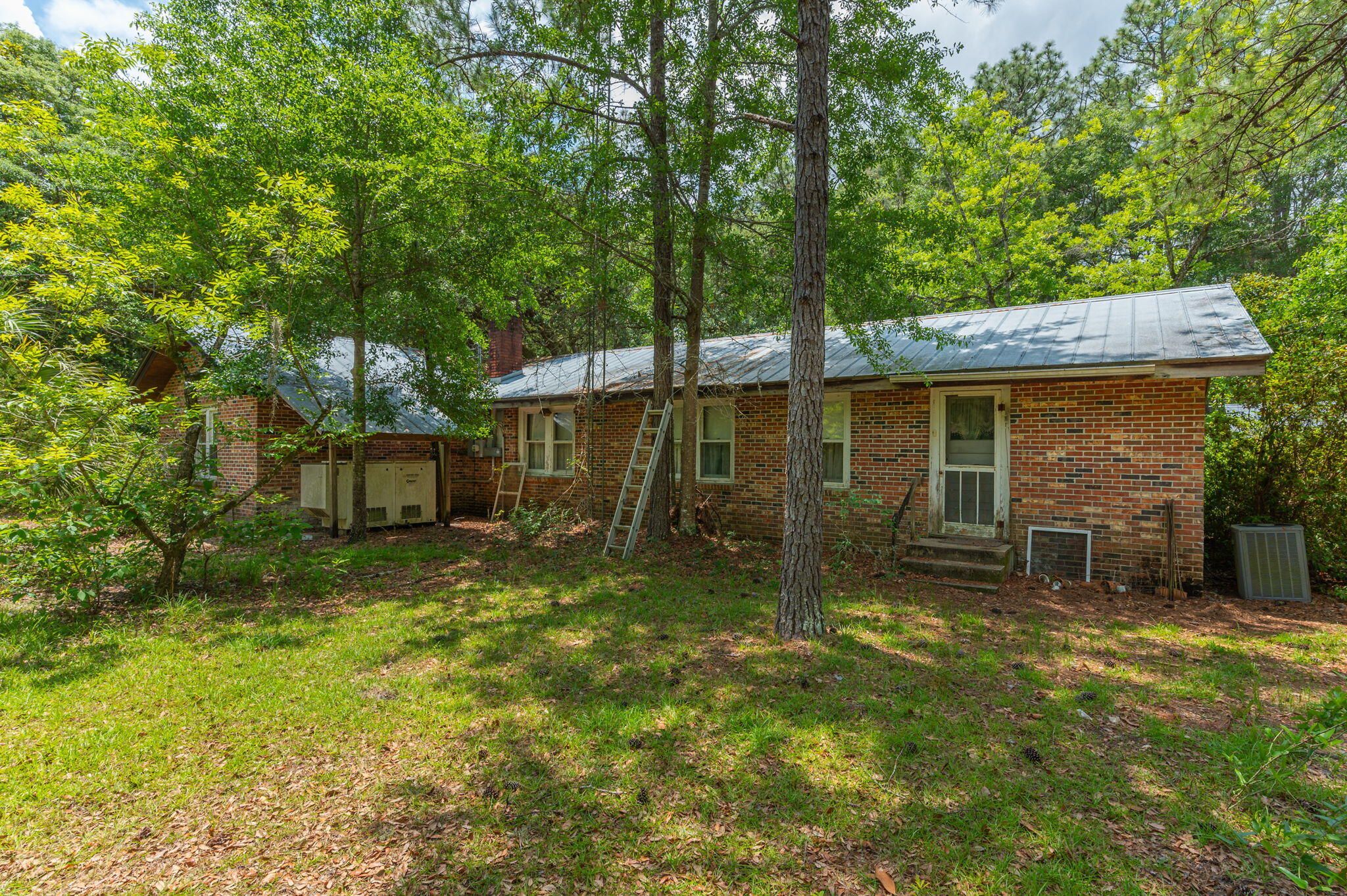 Red brick house with a metal roof, surrounded by trees, with a ladder leaning against the wall.