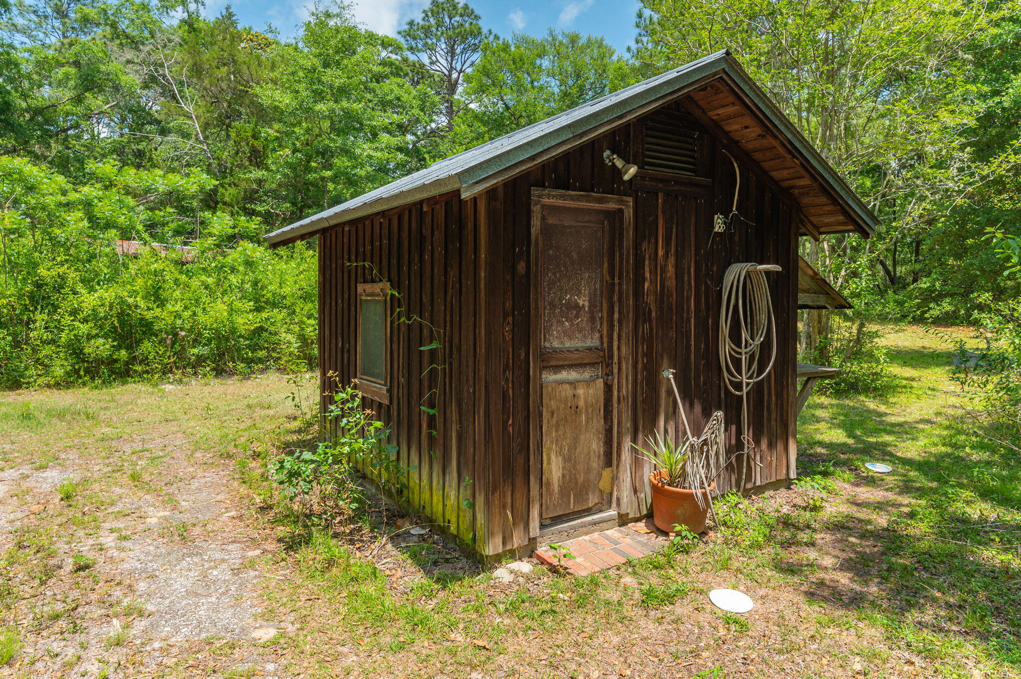 Weathered wooden outbuilding with a gabled roof, coiled hose, and potted plant in a wooded yard.
