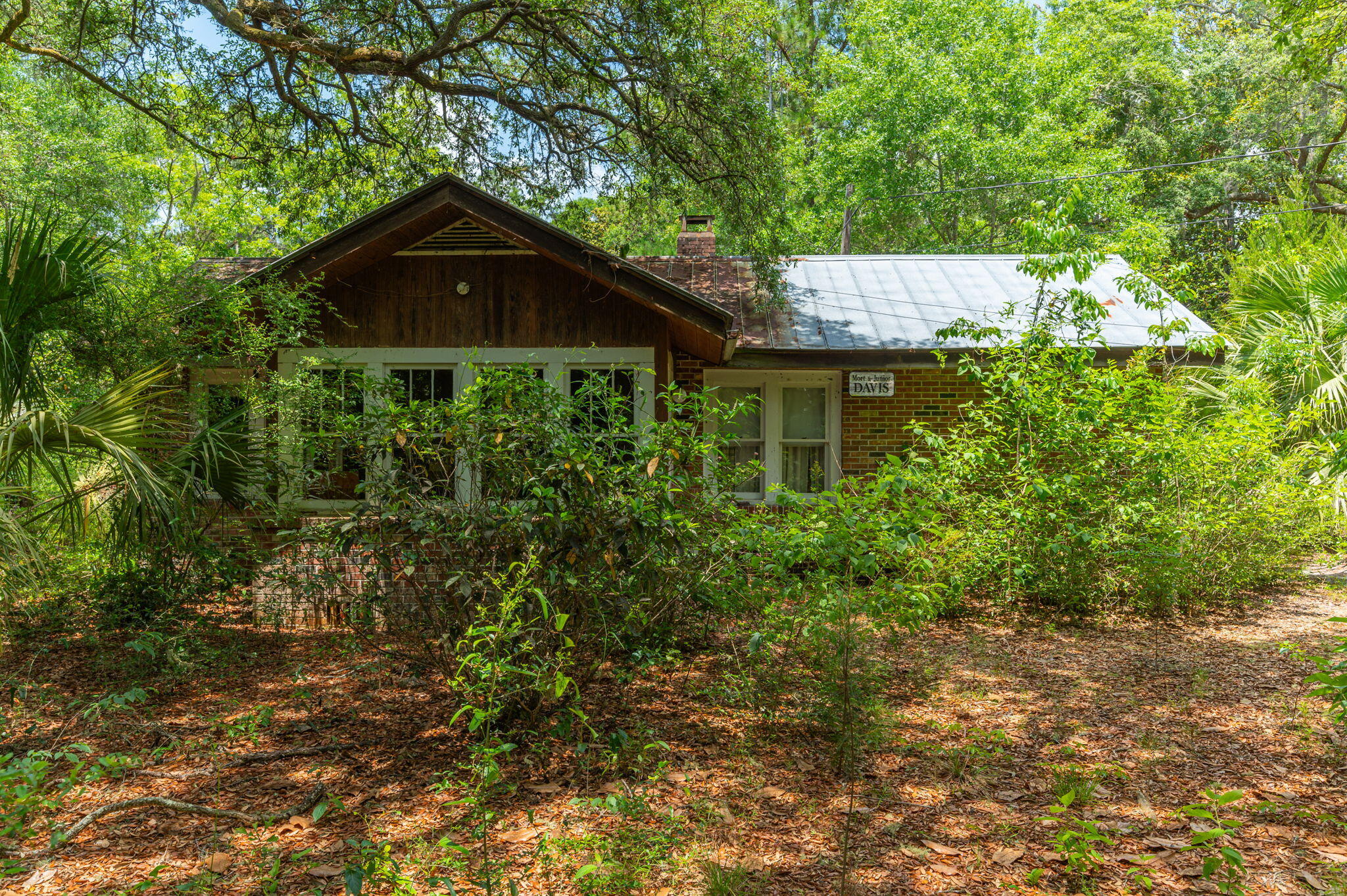 Wood-and-brick house with a metal roof, partly screened by dense woodland and shrubs.