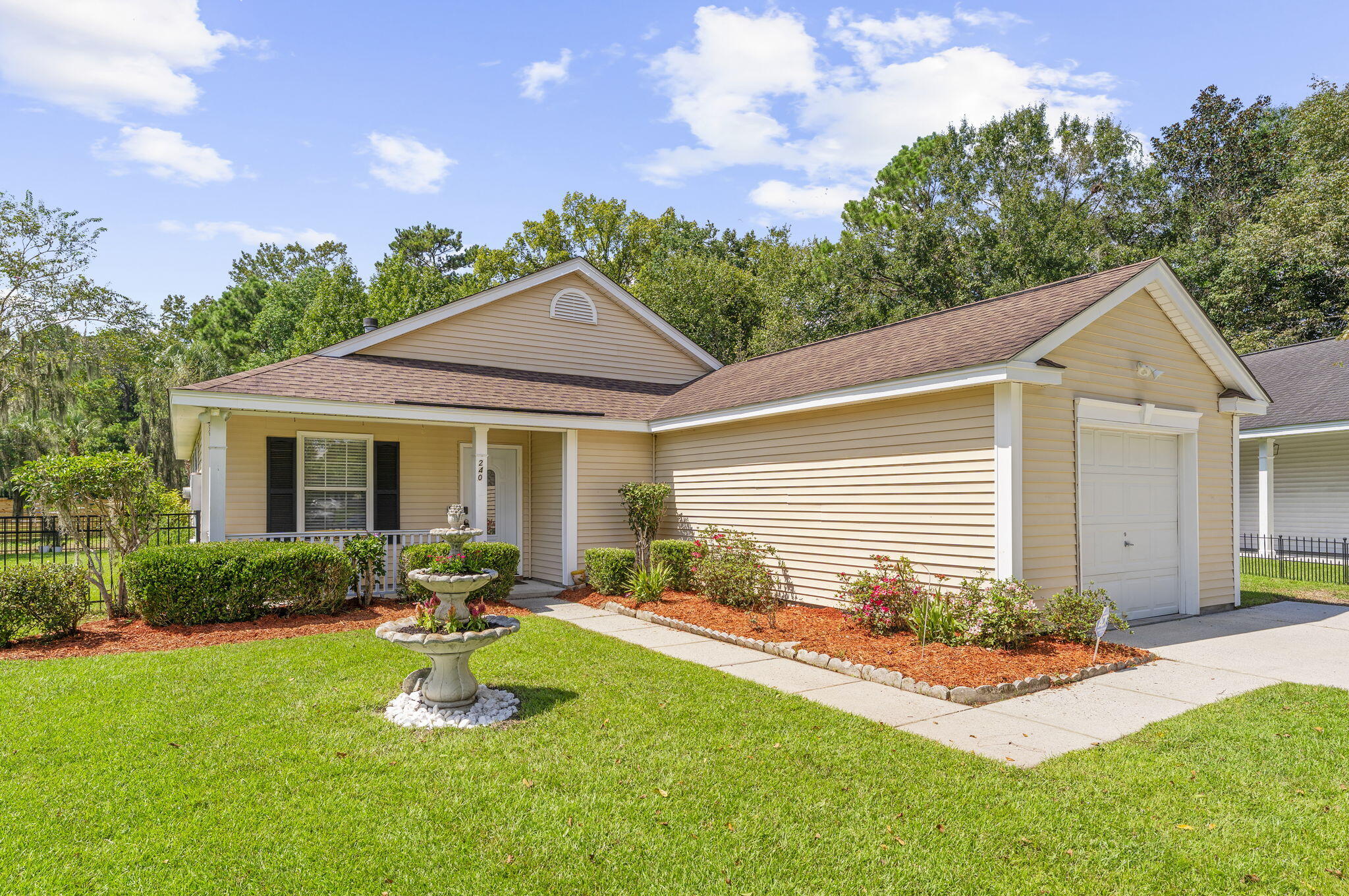 Beige single-story home with an attached garage, covered porch, landscaped beds, and a tiered fountain in the lawn.