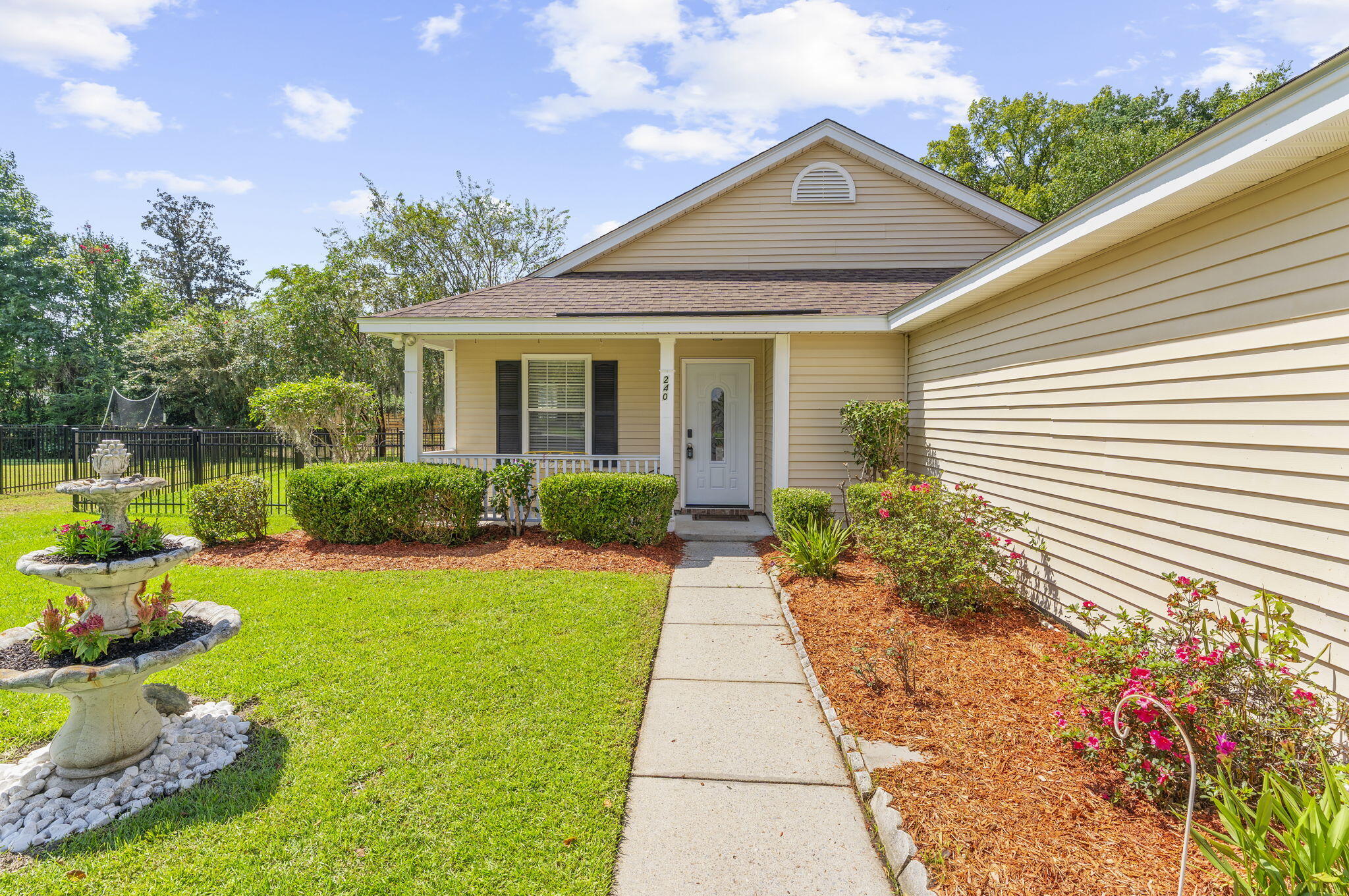 Beige-sided home with a covered porch, landscaped walkway, tiered fountain, and fenced lawn.