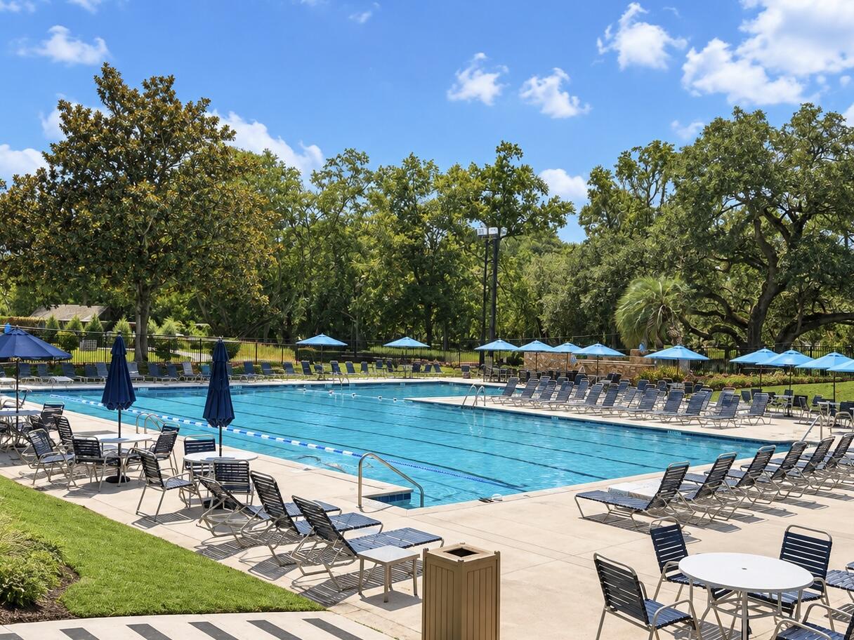 Outdoor pool surrounded by lounge chairs and blue umbrellas beneath mature trees.