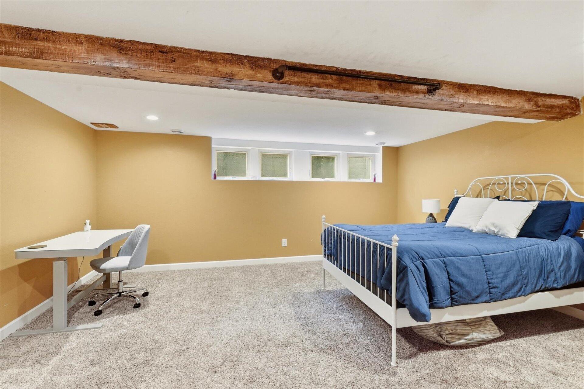 Bedroom with a blue-covered metal bed, desk, high windows, and exposed wood ceiling beam.