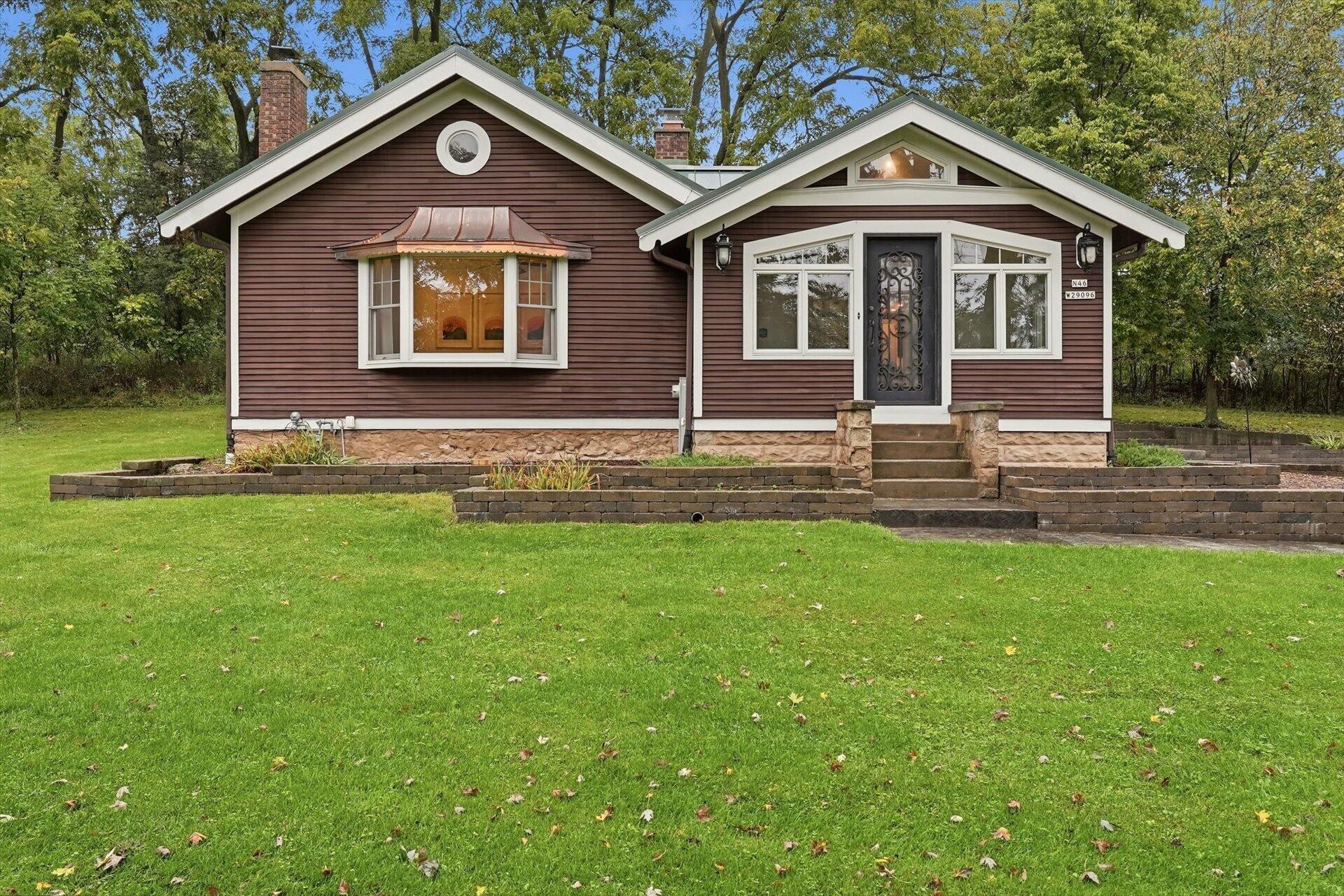 Brown house with white trim, a bay window, decorative entry door, and tiered brick garden walls.