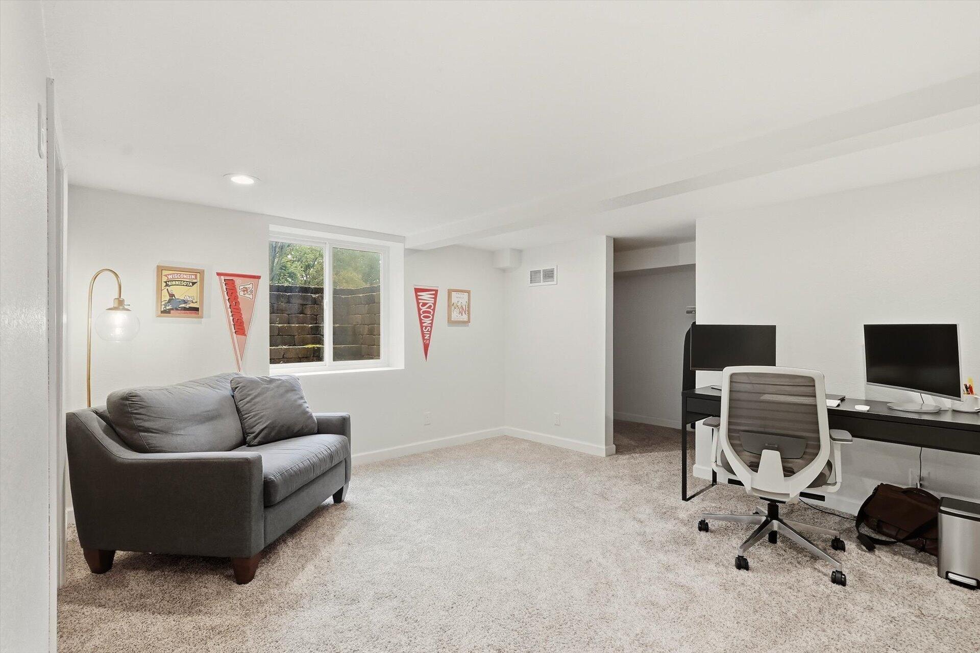 Basement office and sitting area with a gray sofa, desk, two monitors, and Wisconsin pennants.