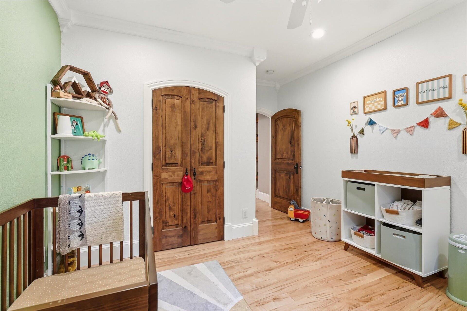 Nursery with a wooden crib, arched wood doors, light flooring, and children’s toy storage.