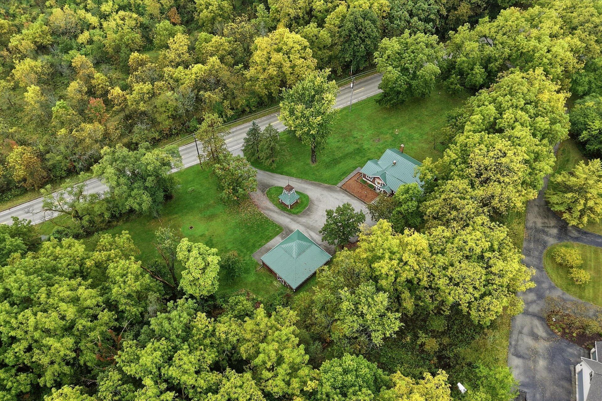 Aerial view of a wooded property with green-roofed buildings and a circular driveway beside a road.