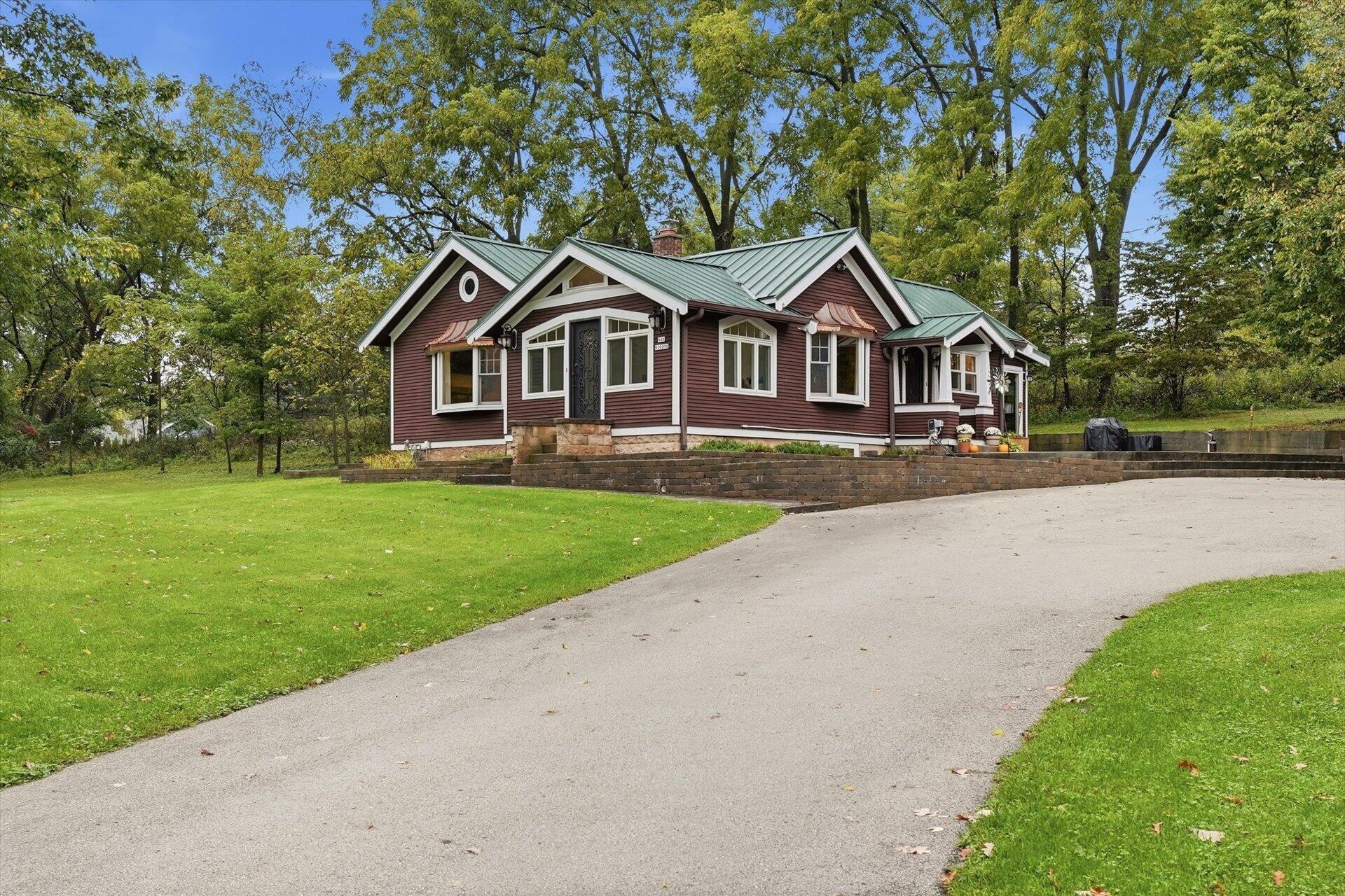 Red-sided single-story home with a green metal roof, mature trees, and a broad paved driveway.