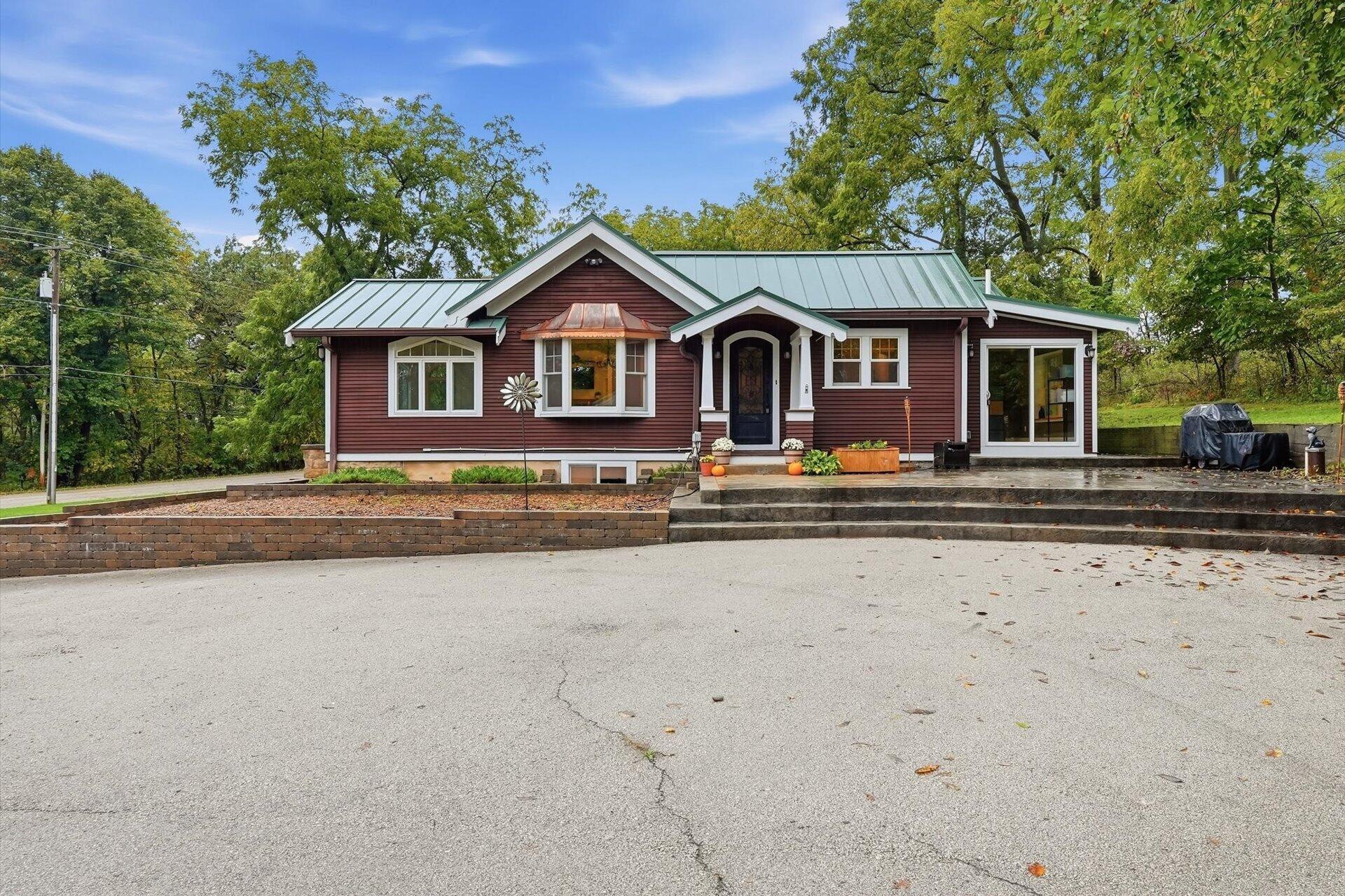 Red-sided home with a green metal roof, covered entry, paved driveway, and mature trees.
