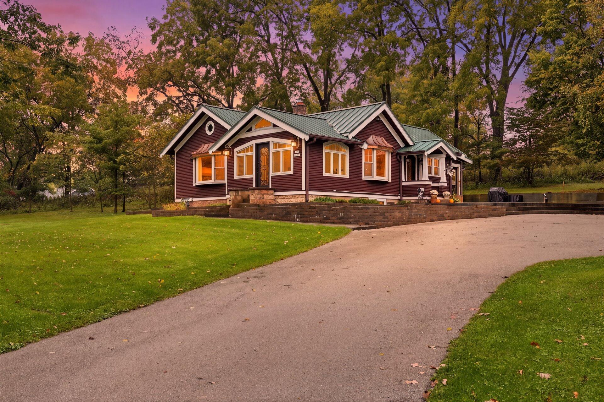 Burgundy home with a green metal roof, lit windows, and a curved driveway at dusk.