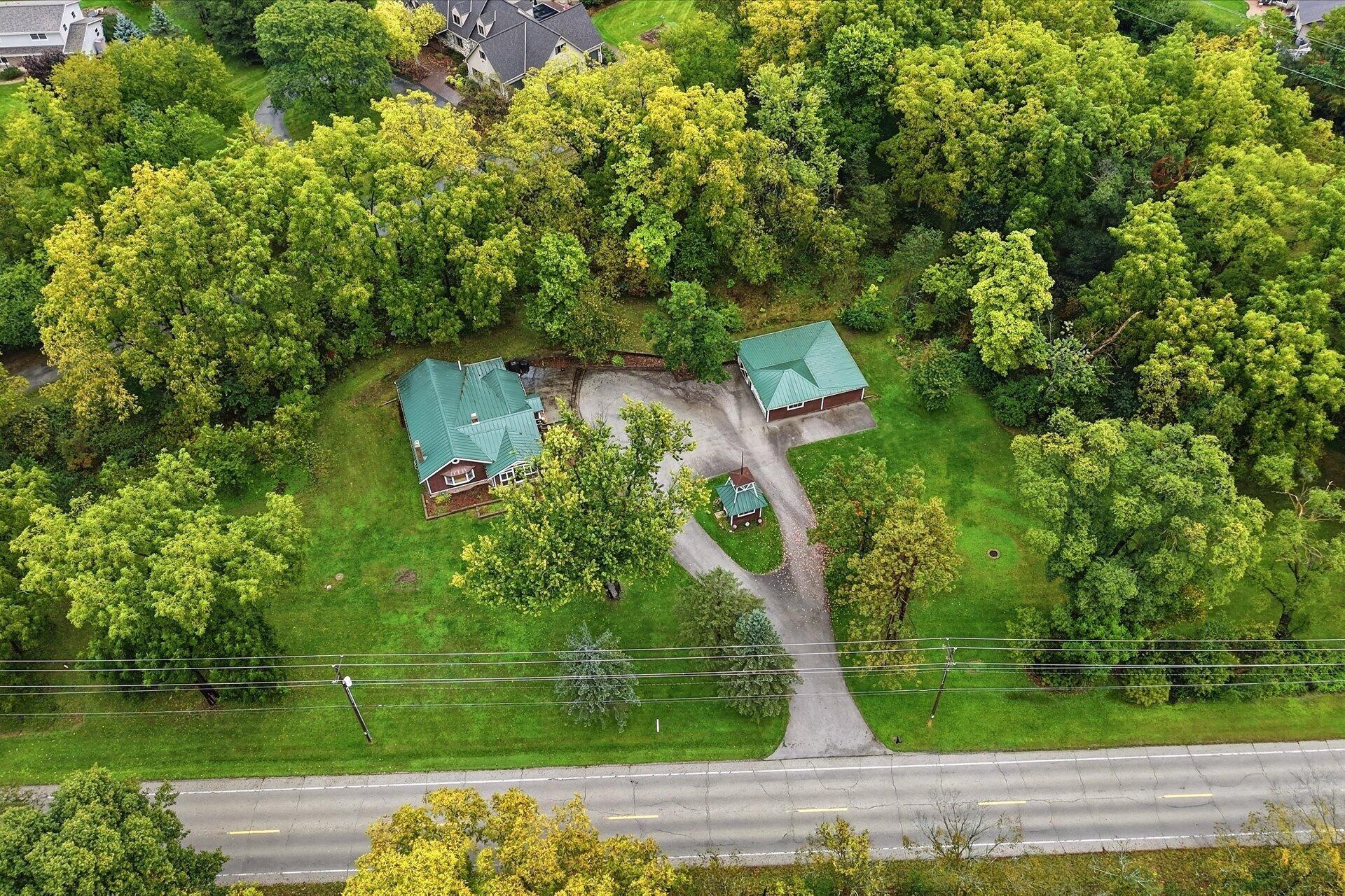 Aerial view of a wooded property with two green-roofed buildings and a driveway to the road.