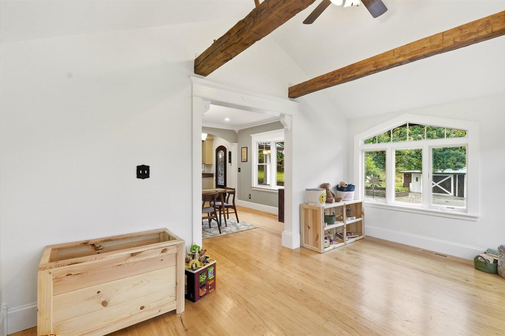 Bright playroom with exposed wood beams, hardwood floors, and large windows overlooking a small outbuilding.