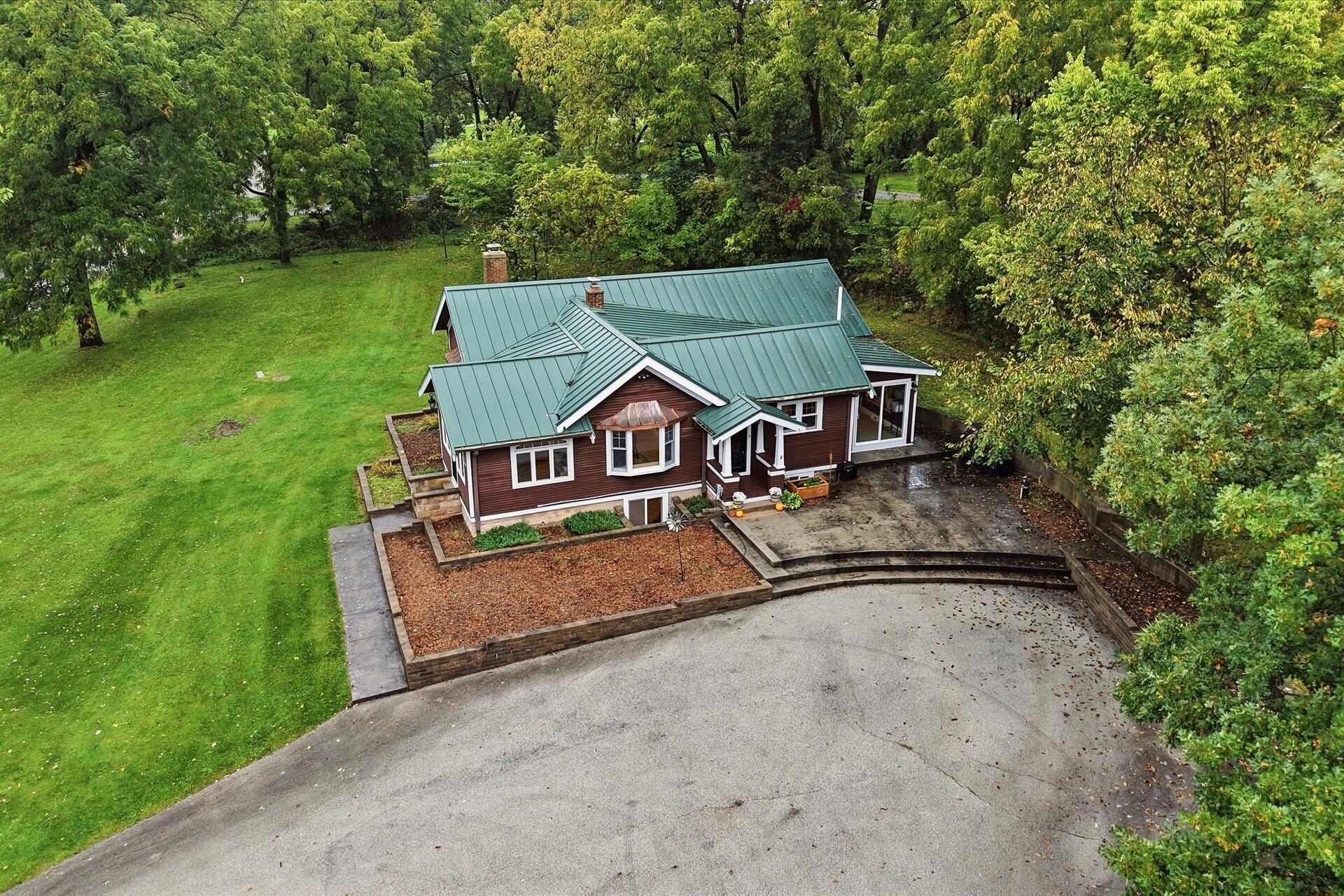 Aerial view of a brown home with a green metal roof, lawn, and mature trees.