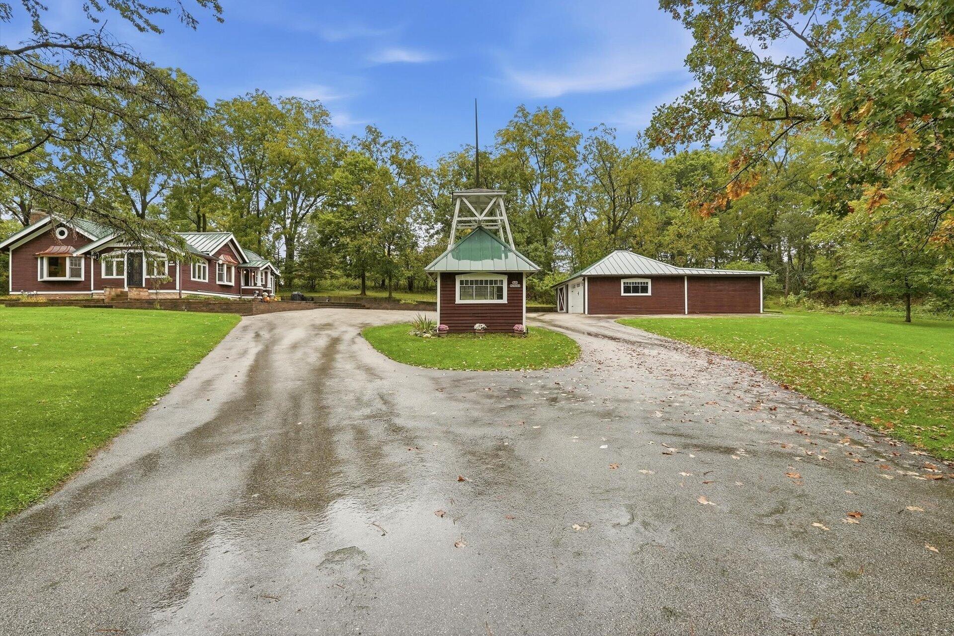 Red-brown house and detached garage framed by trees, with a broad circular driveway and small gate structure.