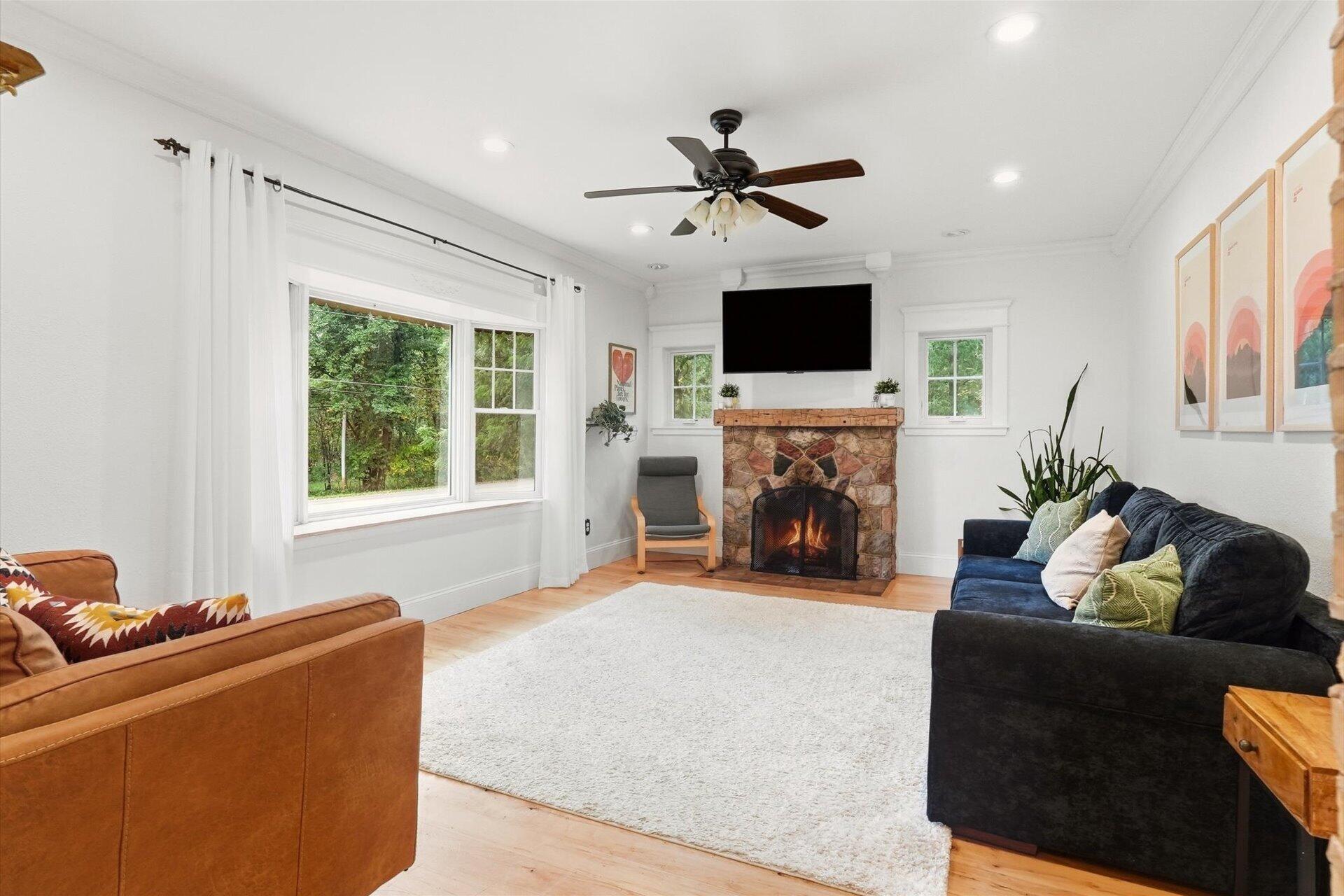 Living room with a stone fireplace, colorful sofas, and large windows overlooking trees.