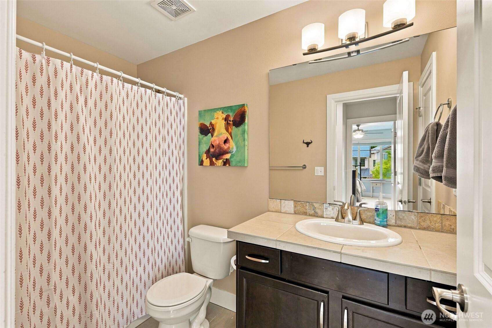 Bathroom with patterned shower curtain, dark vanity, large mirror, and colorful cow painting.