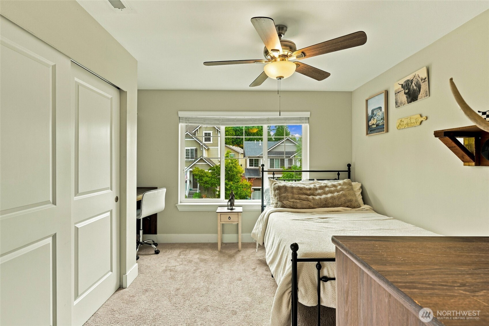 Bedroom with cream bedding, a black metal bed, ceiling fan, and window overlooking neighboring homes.